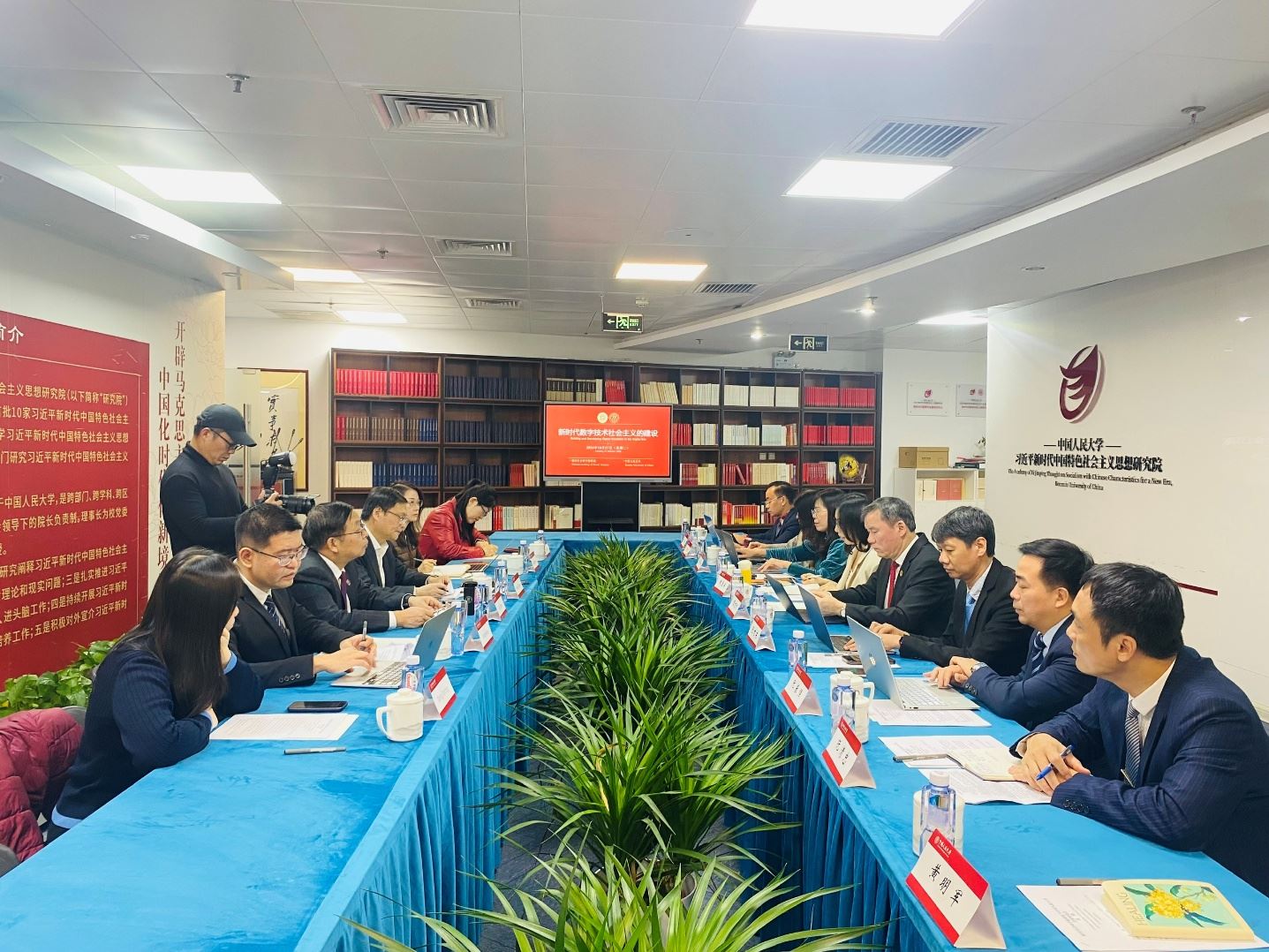 Description: The image depicts a conference room setting with a long table, neatly arranged desks, and chairs, where attendees are seated, engaging in a discussion or presentation. The room is well-lit with ceiling lights and decorations, including red banners and posters.
AI-generated content may be incorrect.