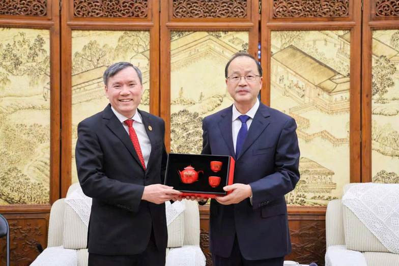 Two men in formal attire stand in front of a decorated red and white ceramic vase.
AI-generated content may be incorrect.